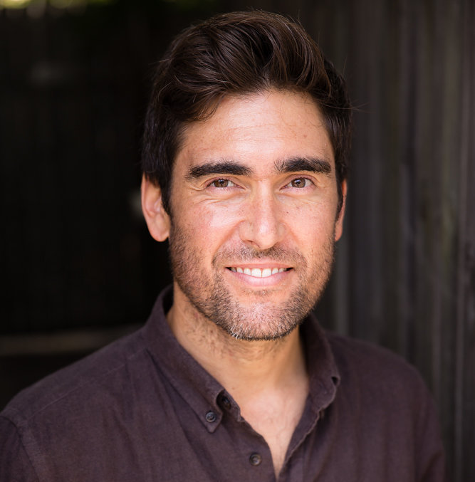 Close-up portrait of a smiling man with short brown hair and light stubble wearing a button-down shirt